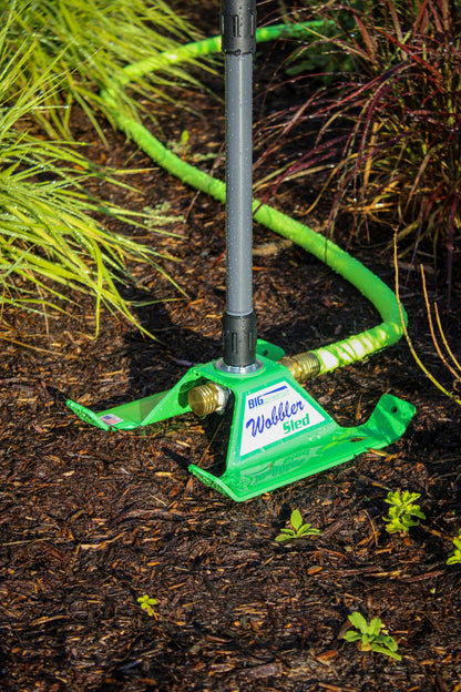 A green Wilden Stord garden hose anchor with a gray metal pole inserted into it, set in a garden bed on dark soil with mulch and green plants.