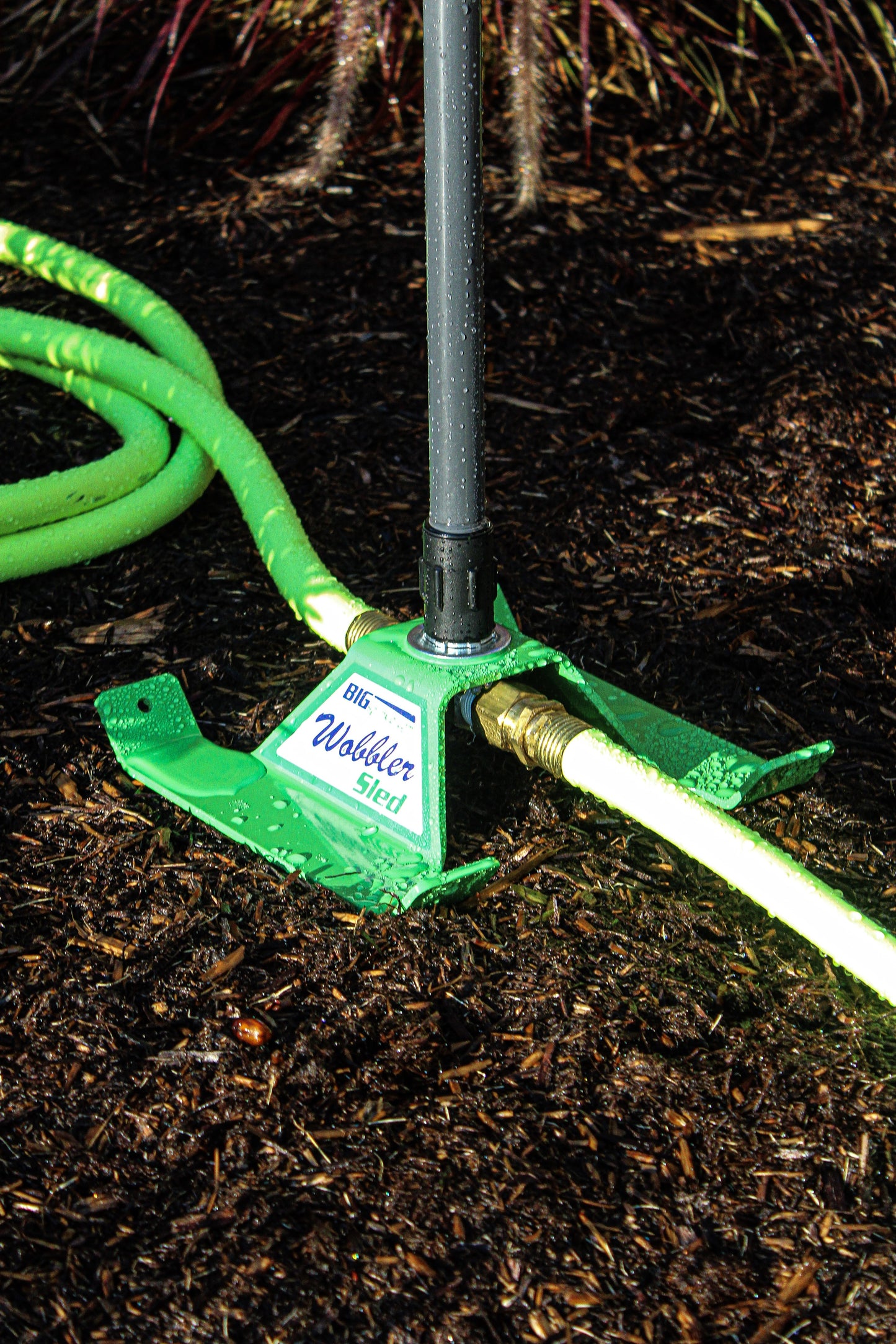 A green Wildebird Watering Hoses garden hose spigot set in dark mulch, with a bright green garden hose connected to the brass fitting on the spigot base.