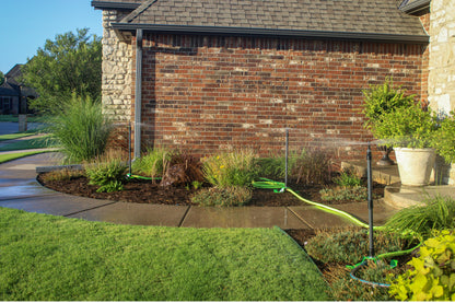 A green garden hose spraying water in a landscaped front yard beside a red brick house, with grass, shrubs, mulch beds, and a stone wall visible.