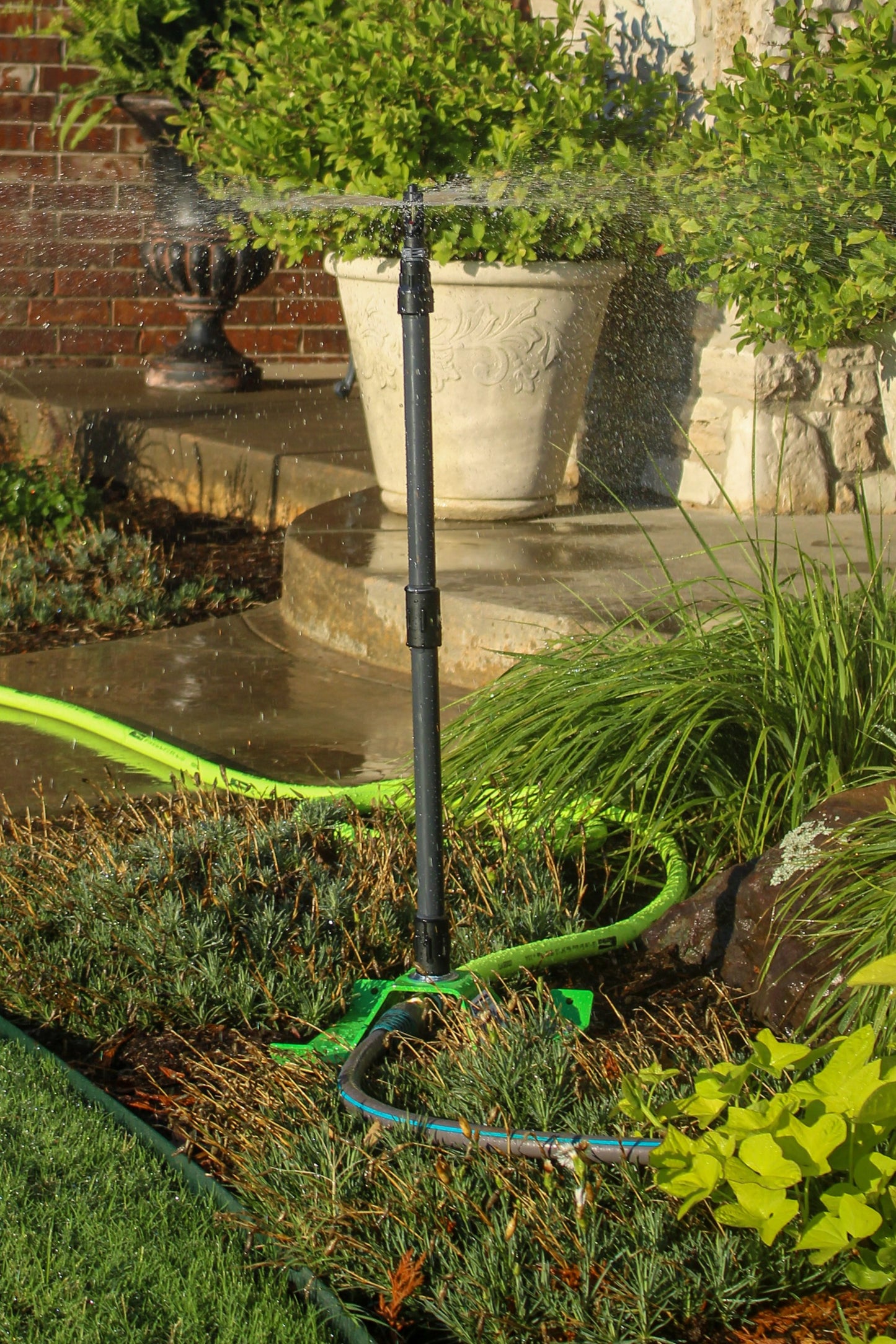 A black automatic sprinkler head spraying water in a landscaped garden bed, connected to a green garden hose. The scene includes potted green plants in large white pots, a brick wall, and stone steps in the background.