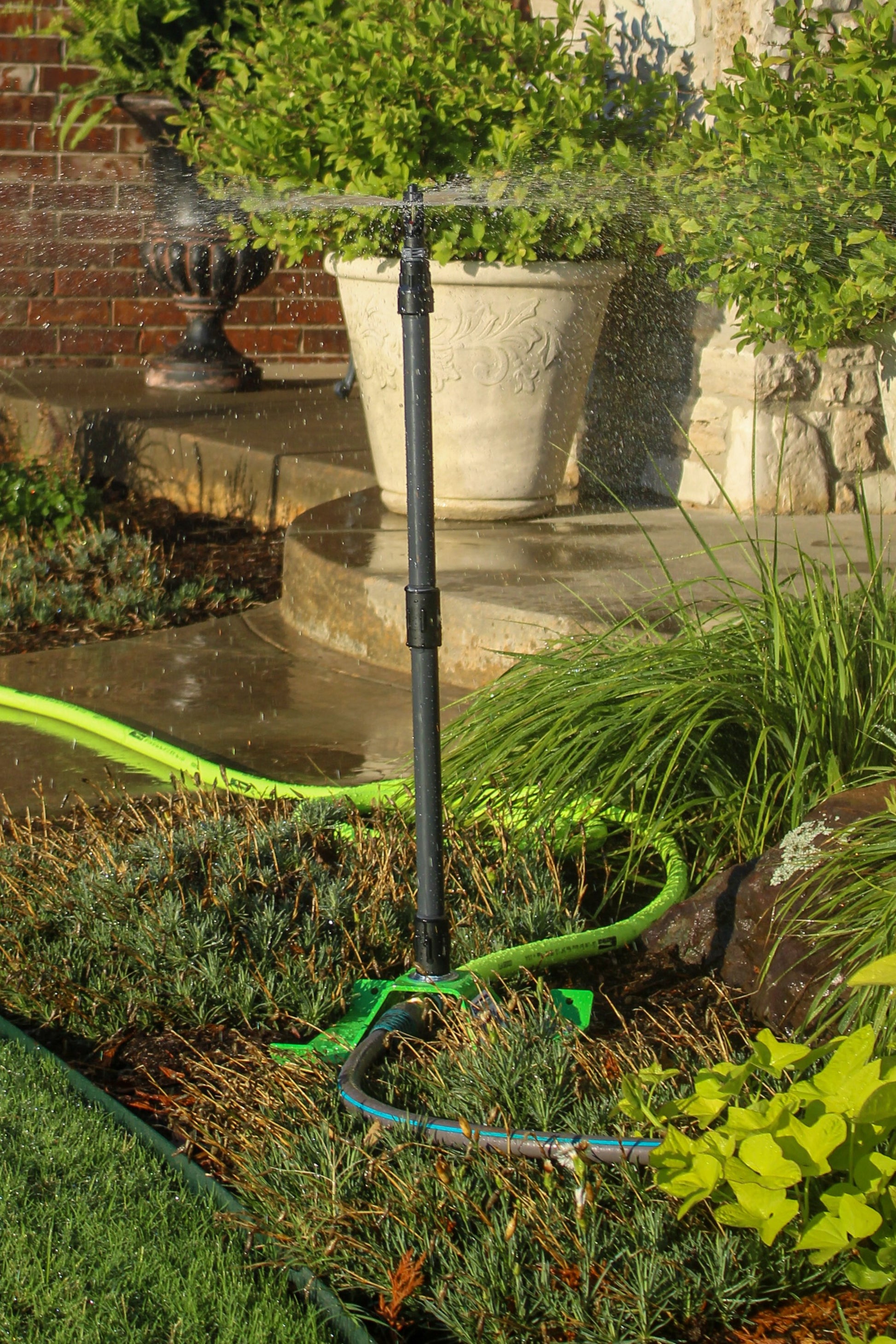 A black automatic sprinkler head spraying water in a landscaped garden bed, connected to a green garden hose. The scene includes potted green plants in large white pots, a brick wall, and stone steps in the background.