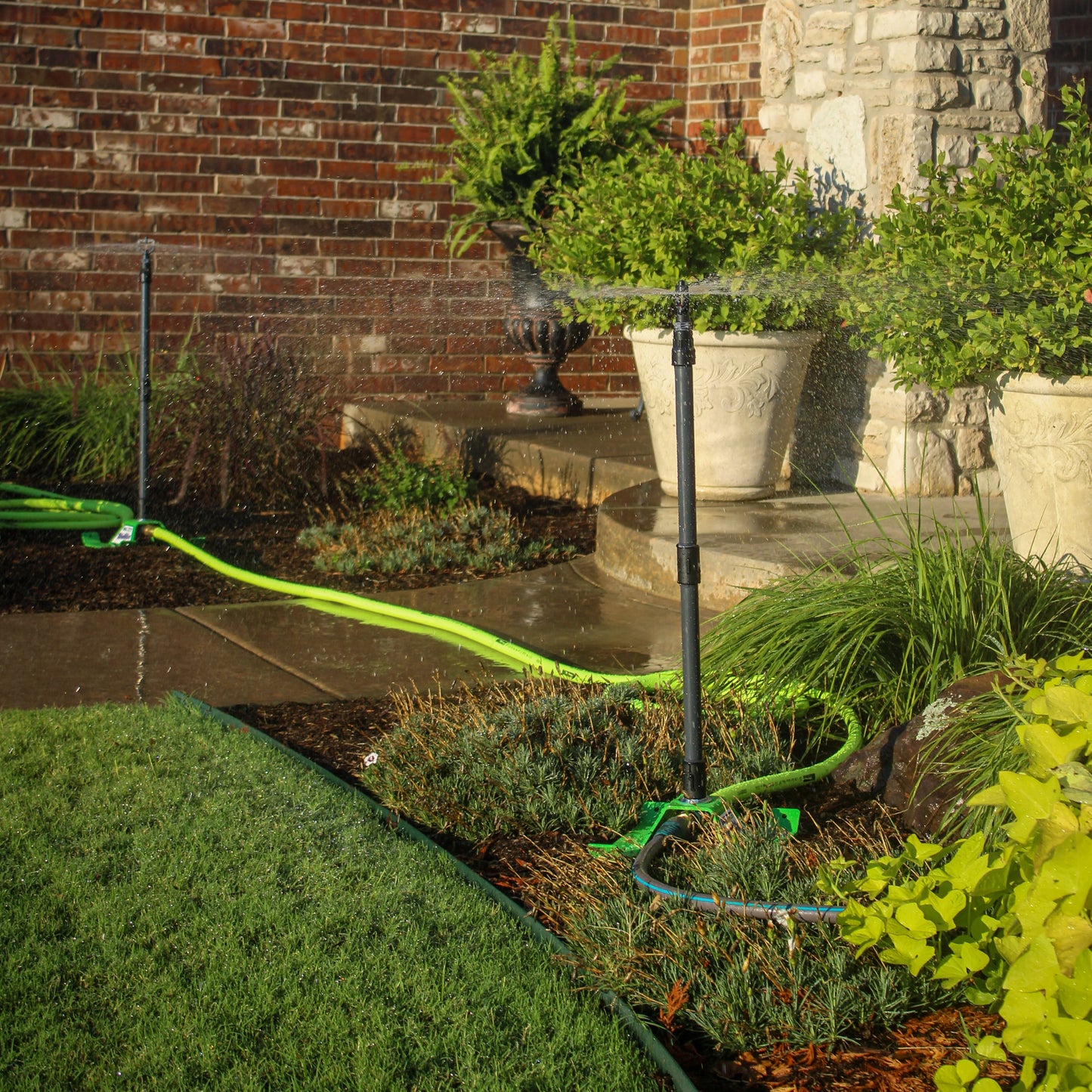 A garden sprinkler system in use, with a bright green hose winding across a landscaped garden bed in front of a brick and stone wall. The sprinkler head is spraying water, wetting the plants and stone steps.