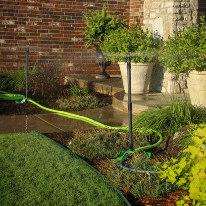 A garden sprinkler system in use, with a bright green hose winding across a landscaped garden bed in front of a brick and stone wall. The sprinkler head is spraying water, wetting the plants and stone steps.