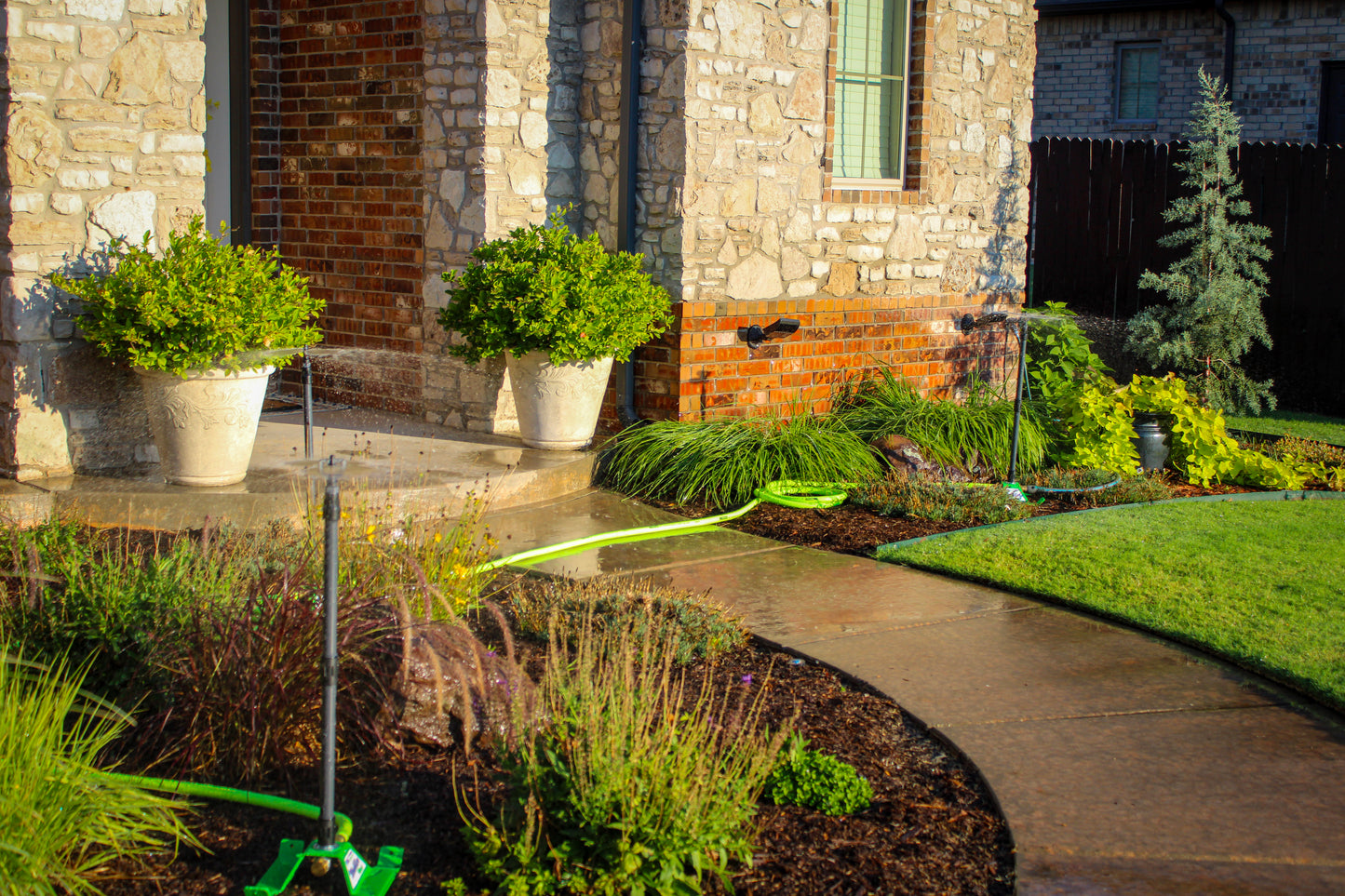 A landscaped front yard beside a stone house, with a curved concrete walkway, green lawn, potted shrubs, and a bright green garden hose laid across the beds.