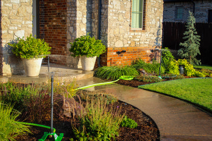 A landscaped front yard beside a stone house, with a curved concrete walkway, green lawn, potted shrubs, and a bright green garden hose laid across the beds.