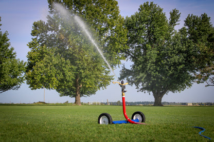 A red and blue wheeled sprinkler watering a green lawn in front of large trees under a blue sky.