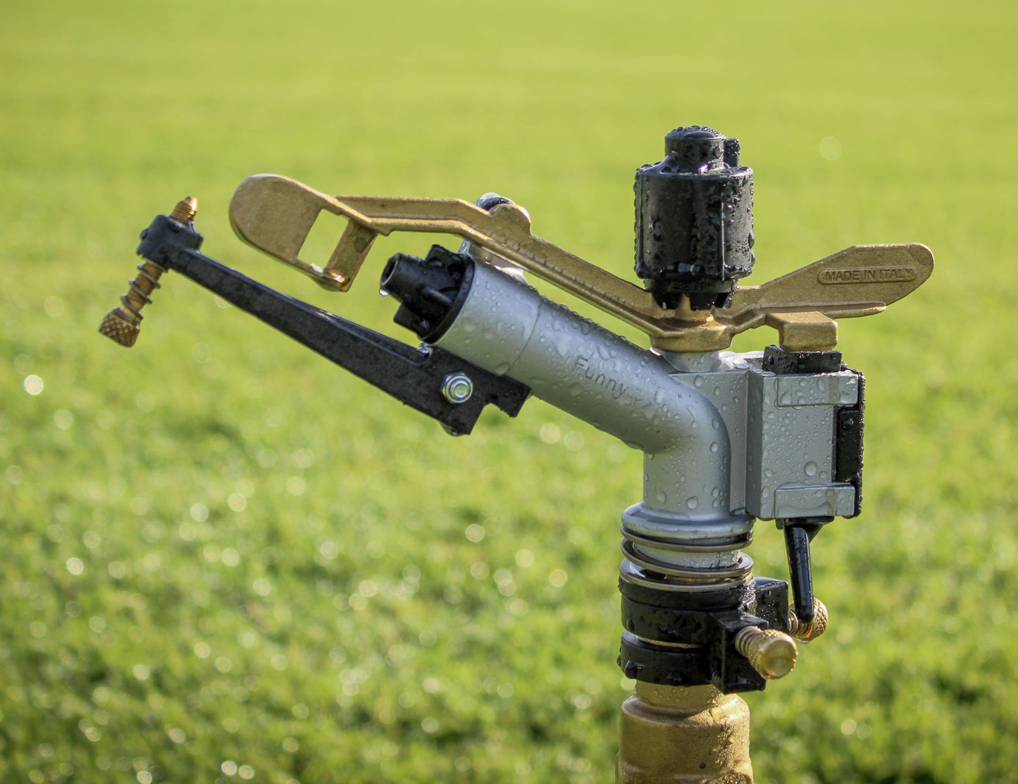 A close-up of a sprinkler head mounted on a post in a grassy field. The sprinkler has a silver body with black and brass components, including a curved arm and top valve assembly.