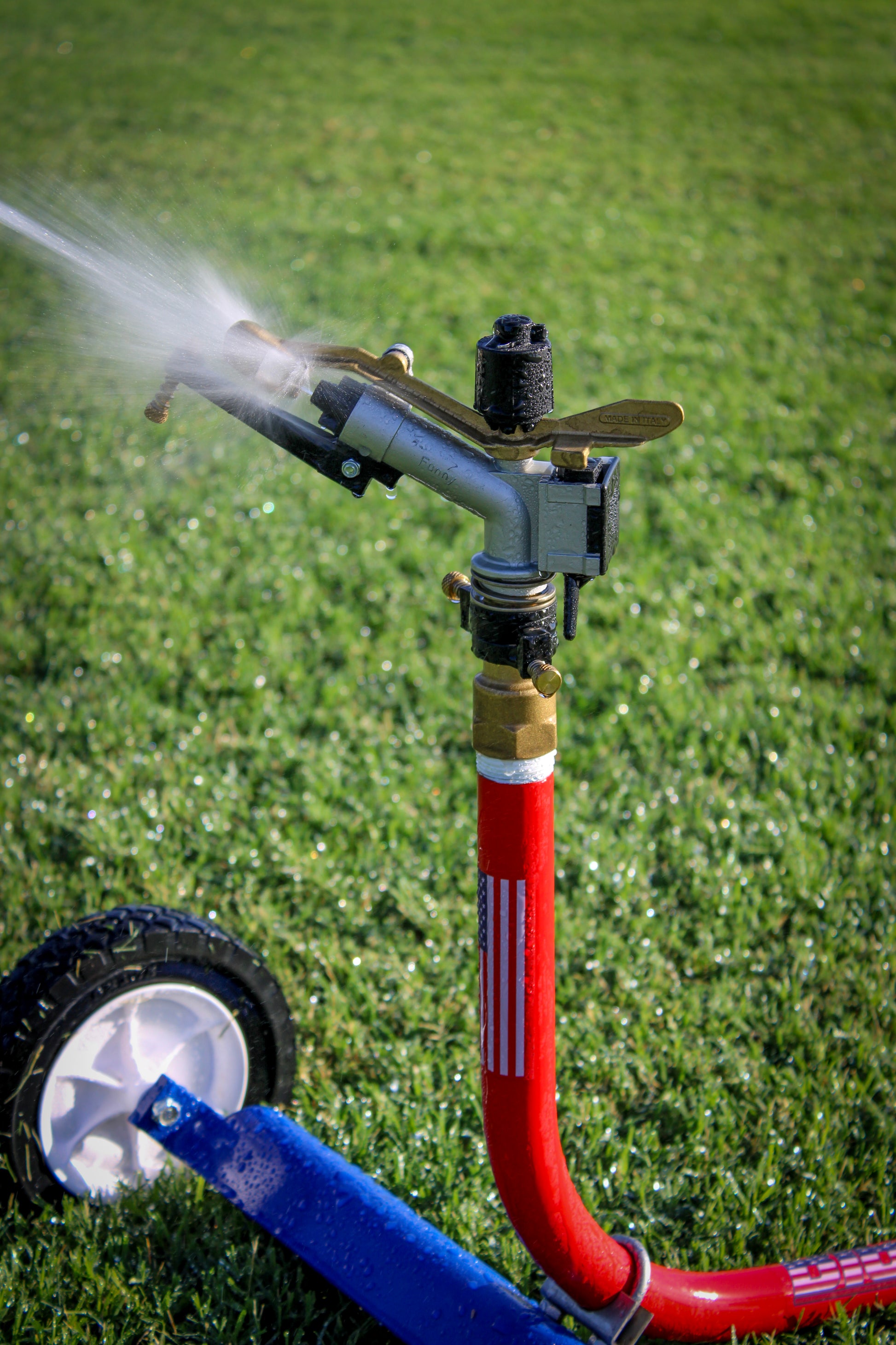 A wheeled garden sprinkler on a green grass lawn, spraying water. The sprinkler has a red hose, a blue handle, a brass and black metal nozzle, and a white wheel.