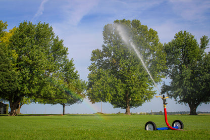 A red and blue wheeled sprinkler watering a large green lawn in a park, with trees, blue sky, and a faint rainbow visible.