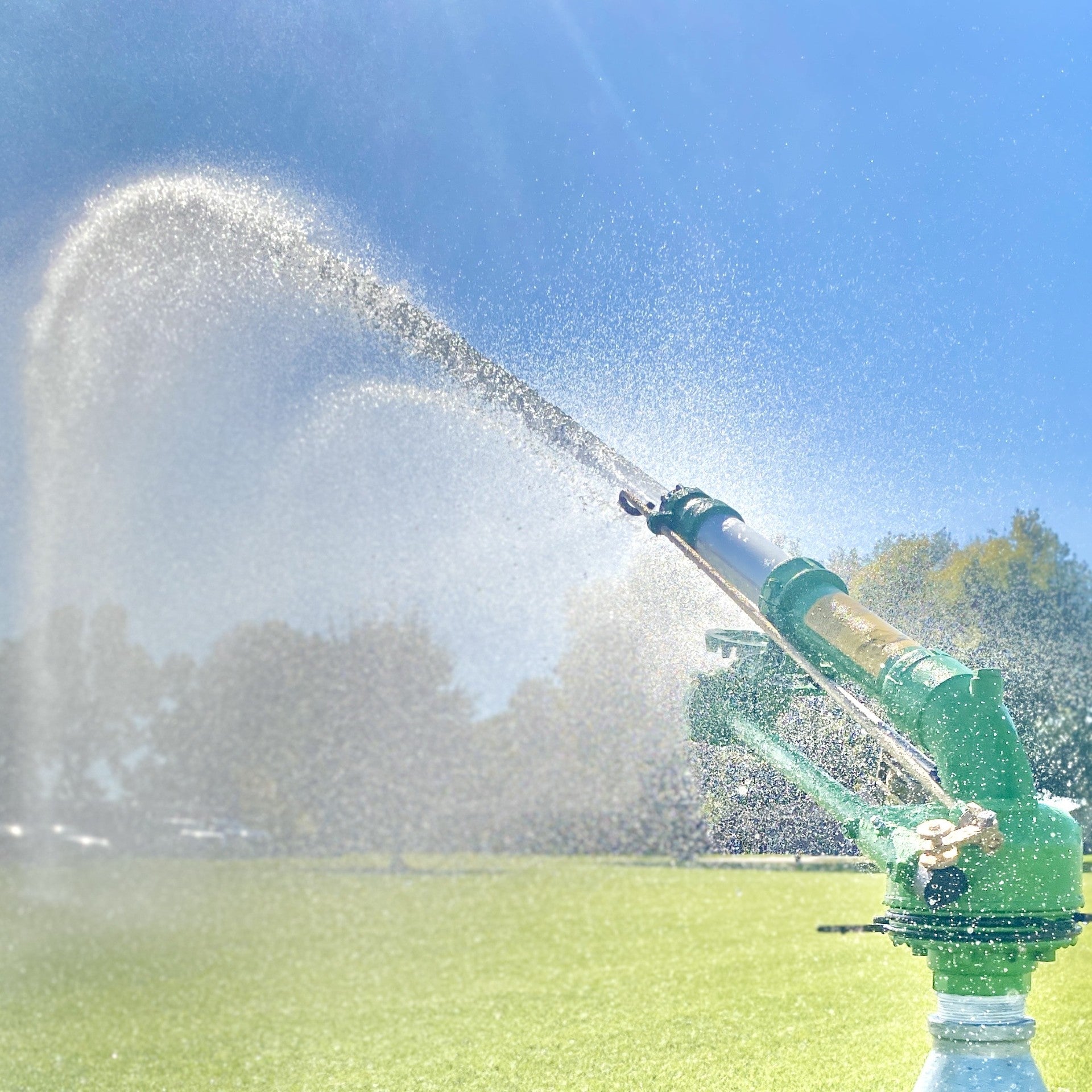 Green Sprinkler is spraying water in an outdoor setting with trees and blue sky.